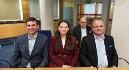 Trzy osoby siedzą w sali konferencyjnej: mężczyzna, kobieta i kolejny mężczyzna, wszyscy skupieni na rozmowie.  