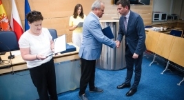 Kobieta trzyma teczkę, dwaj mężczyźni ściskają dłonie przed stołem, symbolizując do składu gremium. 