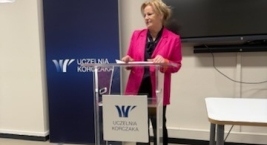 Kobieta przy mównicy, z dużym ekranem w tle, wygłasza przemówienie