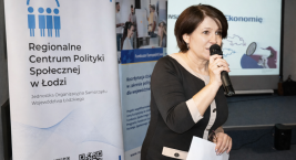 Kobieta przemawia do uczestników wydarzenia, stojąc obok roll-upu Regionalnego Centrum Polityki Społecznej w Łodzi.